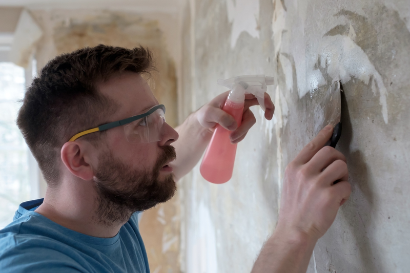 Home repairs. Man removes the old wallpaper from the wall with a spatula and a pulverizer with water.